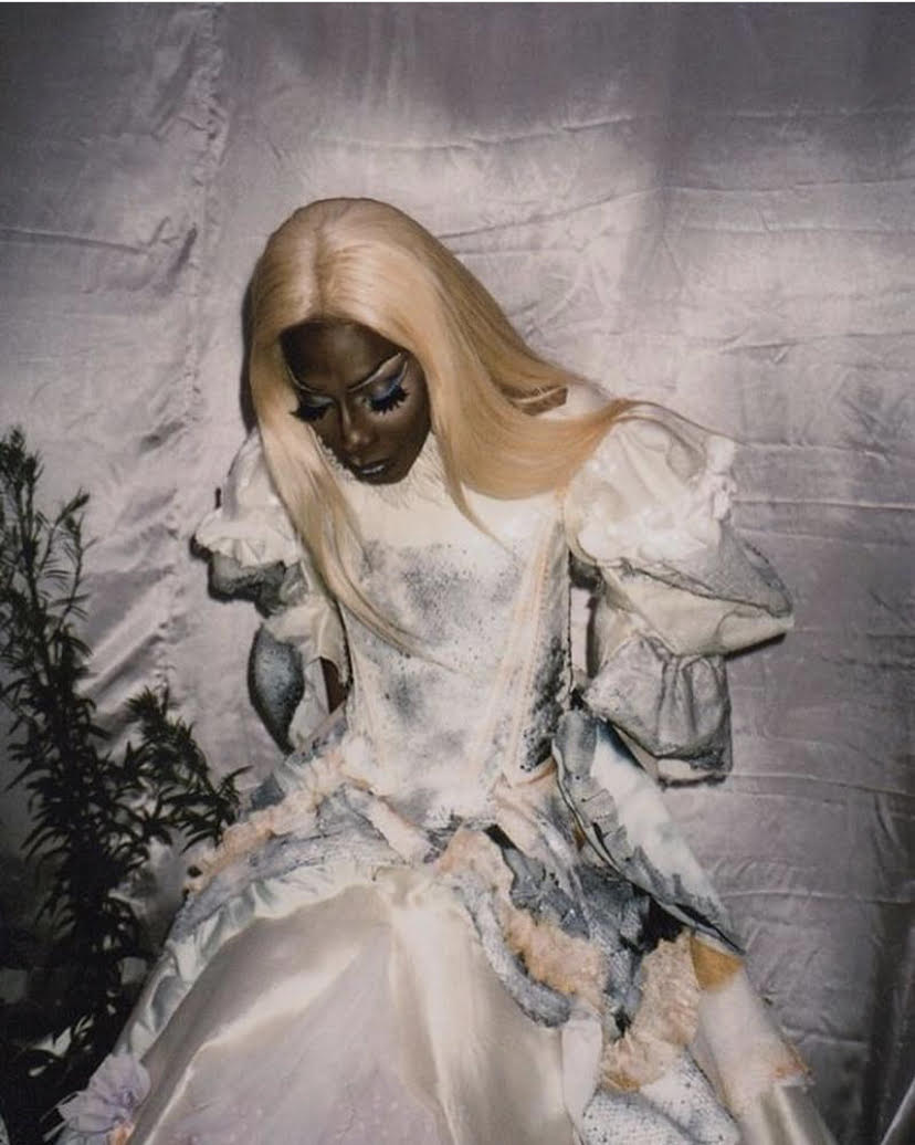 A tall black woman with blond straight hair wears an off=white and grey satin and lace wedding dress. She is seated, with her arms slightly bent, looking vaguely downwards. She has full white and blue eye makeup with long false lashes. Behind her is a white satin back drop, with some greenery to her side.
